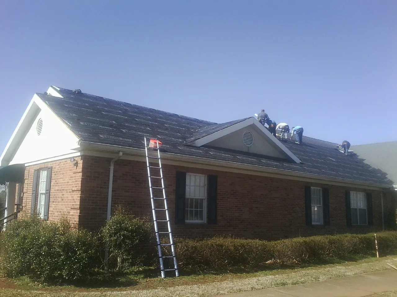 Crew tearing shingles off a brick ranch roof with a ladder set — Emergency Roof Repair work by our crew in Glenburn