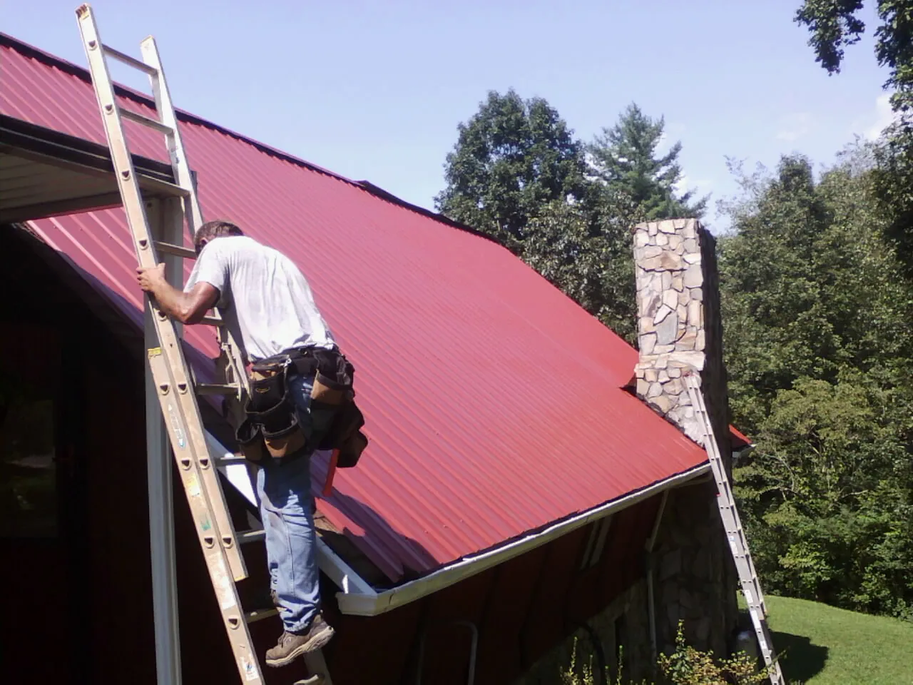 Roofer on a ladder beside a red metal roof and stone chimney — Soffit & Fascia Repair by Roofing Brothers in Glenburn