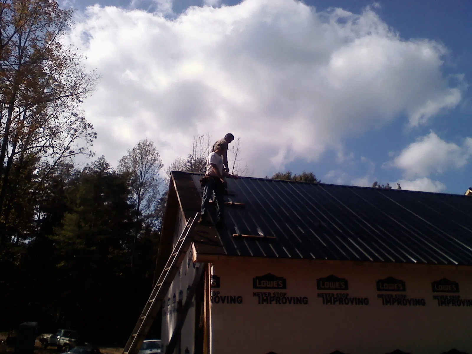 Roofer fastening dark metal panels high against a cloudy sky, from a Roof Inspection job in Glenburn